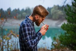 Rauchender bärtiger Mann im Freien in der Nähe des Sees am Abend. schlechte Angewohnheit, Sucht foto