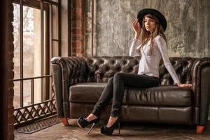 Schönes Mädchen mit schwarzem Hut, das auf dem Sofa sitzt, stilvolle Frau foto