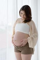 Porträt einer schwangeren Frau, die den Bauch berührt und am Fenster steht foto