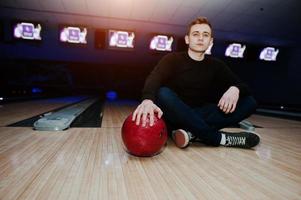 Junger Mann, der eine Bowlingkugel hält, die mit ultraviolettem Licht gegen Bowlingbahnen sitzt. Fokus auf Kugel foto