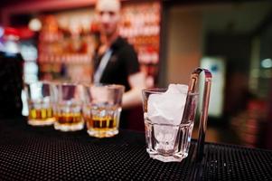 Glas mit Eis und Zange mit drei Whiskygläsern Hintergrund Barmann foto