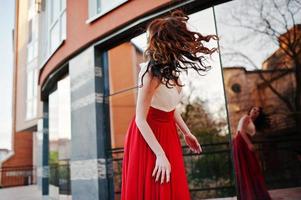 Porträt eines modischen Mädchens im roten Abendkleid mit Haaren in der Luft stellte Hintergrundspiegelfenster des modernen Gebäudes foto