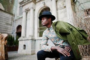 Modeporträt eines schwarzen Afroamerikaners auf grüner Samtjacke an seinen Schultern und schwarzem Hut, sitzend auf gefälltem Baumhintergrund Altes Vintage-Haus mit Bogen und Säulen foto