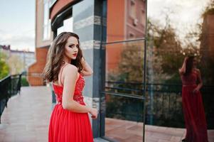 Porträt des modischen Mädchens im roten Abendkleid stellte Hintergrundspiegelfenster des modernen Gebäudes foto