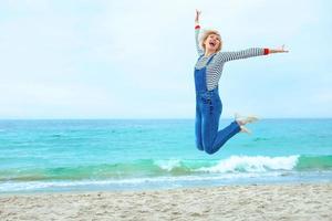 Schöne junge blonde kaukasische Frau im Urlaub in gestreifter Bluse, Turnschuhen und Denim-Overall, die am Strand vor dem erstaunlichen blauen Meereshintergrund springt foto