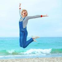 Schöne junge blonde kaukasische Frau im Urlaub in gestreifter Bluse, Turnschuhen und Denim-Overall, die am Strand vor dem erstaunlichen blauen Meereshintergrund springt foto