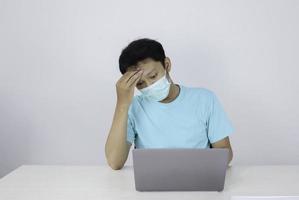 junger asiatischer mann, der eine medizinische maske trägt, fühlt sich ungesund, müde und verwirrt mit der arbeit in laptops auf dem tisch. foto