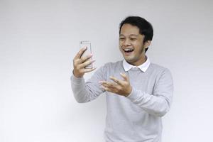 junger asiatischer mann schockiert und glücklich über das, was er auf dem smartphone auf isoliertem grauem hintergrund sieht. foto