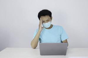 junger asiatischer mann, der eine medizinische maske trägt, fühlt sich ungesund, müde und verwirrt mit der arbeit in laptops auf dem tisch. foto