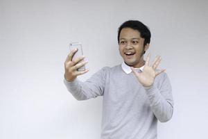 junger asiatischer mann ist glücklich und lächelt, wenn er einen videoanruf tätigt und selfie vor weißem hintergrund macht foto