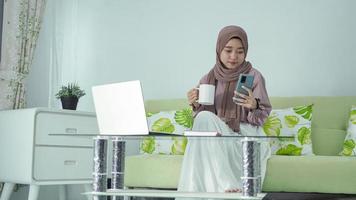 junge Frau im Hijab, die auf ihrem Smartphone nach Inspiration sucht, während sie zu Hause einen Drink genießt foto