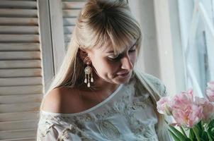 blonde Frau mit Blumen am Fenster, sanftes Lächeln foto