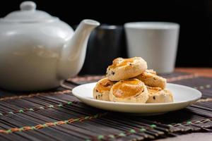 Cashewnuss- und schwarze Seasam-Kekse auf weißem Teller und Holztisch mit verschwommener Kanne Tee auf schwarzem Hintergrund. Nachmittagstee-Konzept. foto