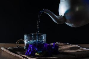 Gießen Sie Schmetterlingserbsentee mit Metalltopf in eine Tasse mit violetter Blume auf braunem Tischtuch auf schwarzem Hintergrund. gesundes getränk zum trinken. kräuter und medizinisches konzept. foto