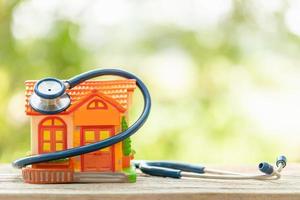 Hand, die blaues Stethoskop und orangefarbenes Hausmodell auf Holztisch hält. Haus- und Gebäudeprüfung foto