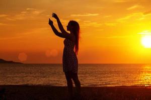 weibliche Silhouette bei Sonnenuntergang am Strand, Hände hoch foto