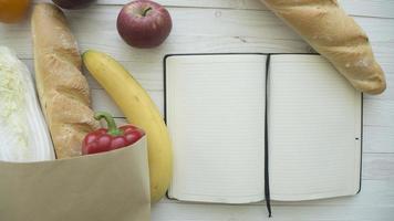 volle papiertüte mit lebensmitteln mit leerem kleinen notizbuch auf holztisch, draufsicht foto