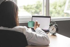 Morgenkaffee im Büro. Rückansicht einer Geschäftsfrau, die im Büro vor einem Laptop mit leerem Bildschirm sitzt, während sie eine Tasse Kaffee in der Hand hält. Mockup-Bildschirm für Anzeigen. foto