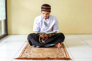 Vorderansicht des jungen asiatischen Muslims halten Gebetsperlen und lesen das heilige Buch al-Quran auf der Gebetsmatte foto