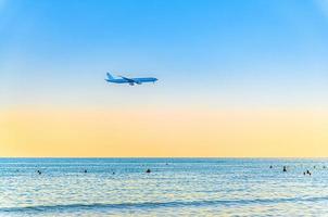 Flugzeug, das tief über dem Meer fliegt, und Touristen, die im Wasser schwimmen, klarer blauer orangefarbener Himmel bei Sonnenuntergang, Flugzeug, das sich auf die Landung vorbereitet foto