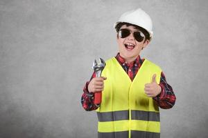 kleiner Kinderbauarbeiter mit weißem Helm und Sonnenbrille und einem Schraubenschlüssel foto