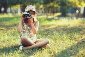 Kleines Mädchen wird von einer Fotokamera gespielt, die auf Gras im Park sitzt. Selfie machen und die Welt um sich herum fotografieren foto