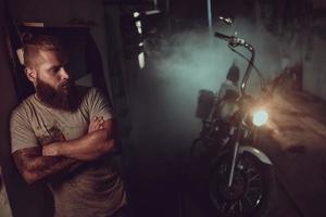 Ein gutaussehender brutaler Mann mit Bart steht in seiner Garage vor dem Hintergrund eines Motorrads und schaut weg foto