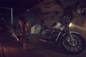Schöner brutaler Mann mit Bart, der ein Motorrad in seiner Garage repariert und mit einer Kreissäge arbeitet. in der garage viele funken und rauch vom sägen foto