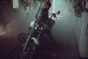 Schöner brutaler Mann mit Bart, der in seiner Garage auf einem Motorrad sitzt, sich die Hände abwischt und zur Seite schaut foto