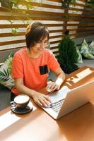 porträt einer selbstbewussten, reifen berufstätigen frau mit brille, einem korallenroten t-shirt, das auf der sommerterrasse im café sitzt, einen laptop für die arbeit benutzt und drinnen glücklich lacht foto