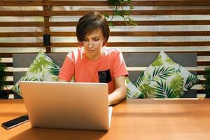 porträt einer selbstbewussten, reifen berufstätigen frau mit brille, einem korallenroten t-shirt, das auf der sommerterrasse im café sitzt, einen laptop für die arbeit benutzt und drinnen glücklich lacht foto