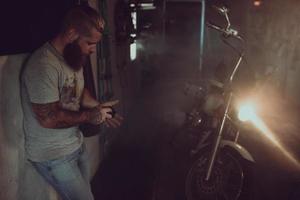 Ein gutaussehender brutaler Mann mit Bart steht in seiner Garage vor dem Hintergrund eines Motorrads und schaut weg foto