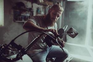 Schöner bärtiger Mann sitzt auf dem Sitz eines Motorrads und repariert das Lenkrad foto