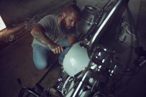 Schöner brutaler Mann mit Bart, der ein Motorrad in seiner Garage repariert