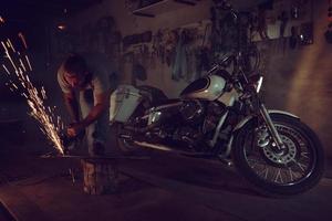 Schöner brutaler Mann mit Bart, der ein Motorrad in seiner Garage repariert und mit einer Kreissäge arbeitet. in der garage viele funken und rauch vom sägen