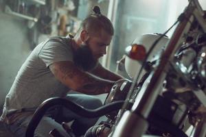 Schöner bärtiger Mann, der sein Motorrad in der Garage repariert. ein Mann in Jeans und T-Shirt foto