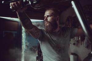 Schöner brutaler Mann mit Bart, der in seiner Garage auf einem Motorrad sitzt und wegschaut foto