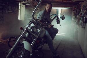 Schöner brutaler Mann mit Bart, der in seiner Garage auf einem Motorrad sitzt, sich die Hände abwischt und zur Seite schaut foto