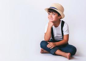 Lächelndes und glückliches Kind mit weißem T-Shirt und Hut, das auf dem Boden auf weißem Hintergrund sitzt foto