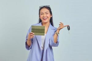 Porträt einer fröhlichen jungen asiatischen Frau, die Fahrzeugbuch zeigt und Fahrzeugschlüssel isoliert auf weißem Hintergrund hält foto