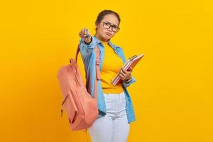 Verärgerte junge asiatische Studentin in Denim-Kleidung mit Rucksack, der Notizbuch hält und Stift auf die Kamera zeigt, isoliert auf gelbem Hintergrund. Bildung im High School University College-Konzept foto