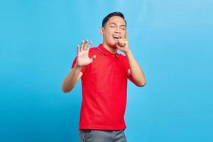Porträt eines attraktiven jungen asiatischen Mannes in rotem Hemd, der laut singt, während er isoliert auf blauem Hintergrund steht foto