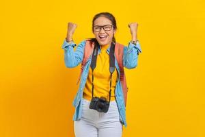 porträt einer aufgeregten jungen reisenden asiatischen frau mit rucksack und kamera in jeanskleidung, während sie das glück einzeln auf gelbem hintergrund feiern. Passagier, der am Wochenende reist. Flugreisekonzept foto