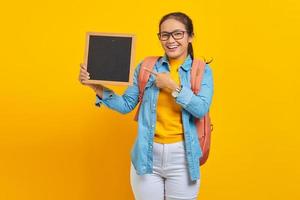 Porträt einer lächelnden jungen asiatischen Studentin in Freizeitkleidung mit Rucksack, die mit dem Finger auf gelbem Hintergrund auf eine leere Tafel zeigt. Bildung im College-Universitätskonzept foto