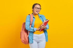 porträt einer fröhlichen jungen asiatischen studentin in lässiger kleidung mit rucksack, der buch hält und isoliert auf gelbem hintergrund in die kamera blickt foto