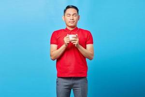 Porträt eines hübschen asiatischen jungen Mannes, der eine Tasse Kaffee hält und seine Augen schließt, während er köstliches Kaffeearoma einatmet, das auf blauem Hintergrund isoliert ist foto