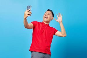 Porträt eines fröhlichen asiatischen jungen Mannes, der einen Videoanruf tätigt und auf die Smartphone-Kamera winkt, die auf blauem Hintergrund isoliert ist foto