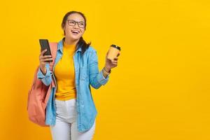 porträt einer fröhlichen jungen asiatischen studentin in jeanskleidung mit rucksack, der tasse kaffee und smartphone isoliert auf gelbem hintergrund hält foto