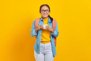porträt einer lächelnden jungen asiatischen studentin in jeanskleidung mit rucksack, der eine tasse kaffee isoliert auf gelbem hintergrund hält. Bildung im High School University College-Konzept foto