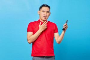 porträt eines überraschten jungen asiatischen mannes, der ein handy benutzt und die kamera isoliert auf blauem hintergrund betrachtet foto